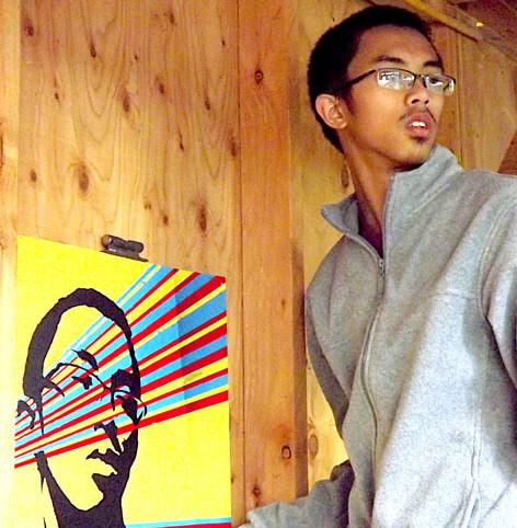 Ty Gocuan poses with his newly created art piece as it is being hung for display. He used a variety of colors and shades to construct his artwork. His piece was chosen as the Artist Statement Piece by members of his class. His portrait is on display with others on Level 3 of the College Complex.